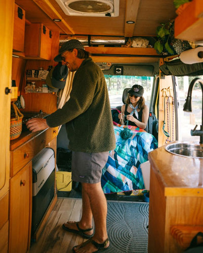 A man using the stove in a camper van while a woman knits in the background, using a Rumpl camping blanket while knitting.